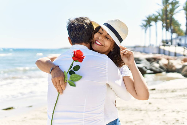 Pareja hispana de mediana edad abrazando y sosteniendo flores en la playa. - Foto, imagen