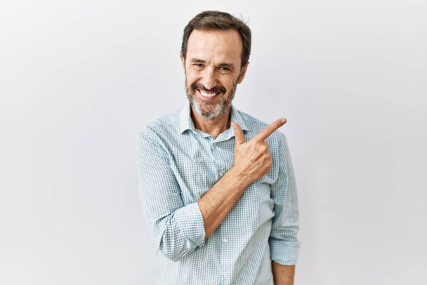 Hombre hispano de mediana edad con barba de pie sobre fondo aislado alegre con una sonrisa en la cara señalando con la mano y el dedo hacia el lado con expresión feliz y natural  - Foto, imagen