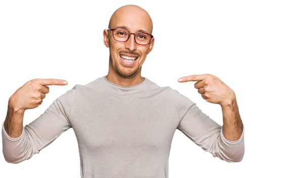 Glatzköpfiger Mann mit Bart, lässiger Kleidung und Brille, der selbstbewusst lächelnd mit den Fingern zeigt, stolz und glücklich.  - Foto, Bild
