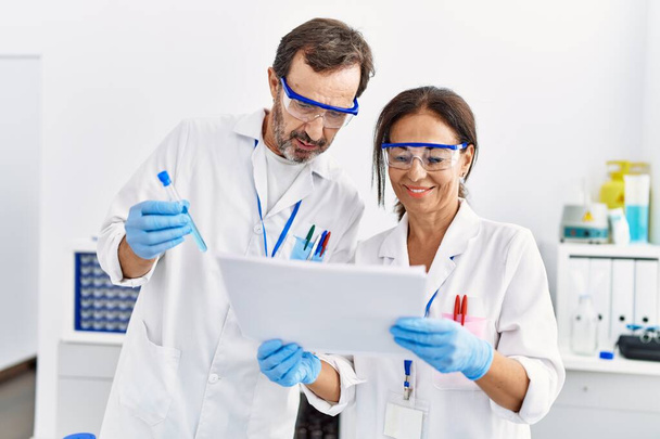 Mann und Frau mittleren Alters tragen einheitliche wissenschaftliche Dokumente und halten Reagenzgläser im Labor - Foto, Bild