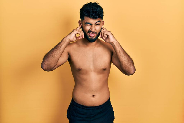 Arabische man met baard draagt zwemkleding zonder shirt over oren met vingers met geïrriteerde uitdrukking voor het lawaai van luide muziek. doof concept.  - Foto, afbeelding