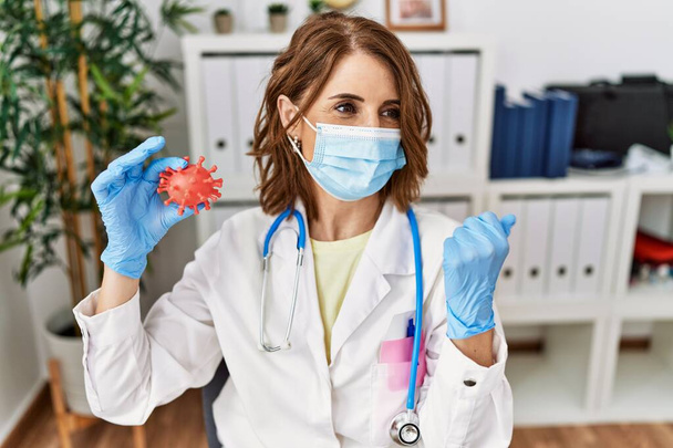 Middle age doctor woman wearing safety mask holding virus toy pointing thumb up to the side smiling happy with open mouth  - Photo, image