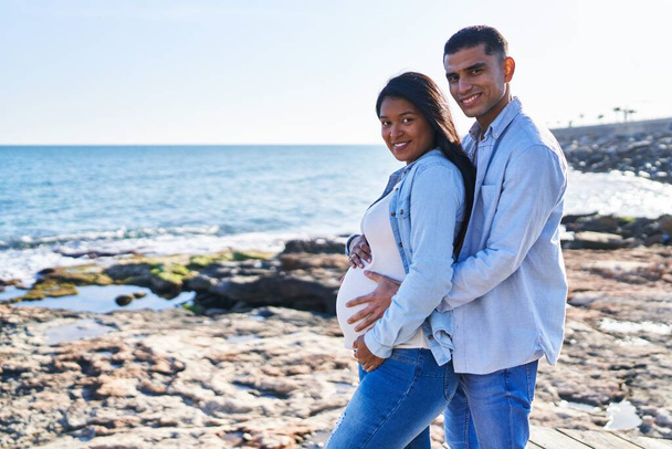 Young latin couple expecting baby hugging each other standing at seaside - Foto, Imagem