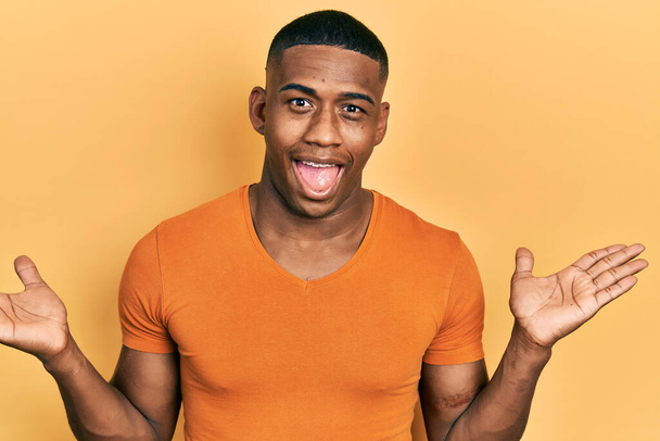 Jonge zwarte man draagt casual oranje t-shirt viert overwinning met vrolijke glimlach en winnaarsuitdrukking met opgeheven handen  - Foto, afbeelding