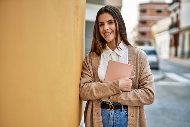 Joven estudiante hispana sonriendo feliz sosteniendo libro en la ciudad. - Foto, Imagen