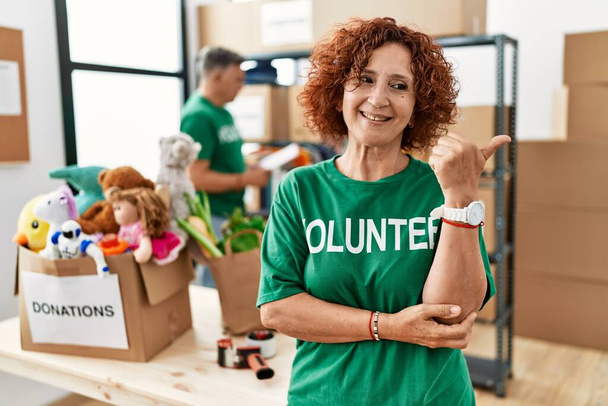 Женщина средних лет в футболке волонтера на пожертвования стоять улыбаясь со счастливым лицом глядя и указывая на сторону с большим пальцем вверх.  - Фото, изображение