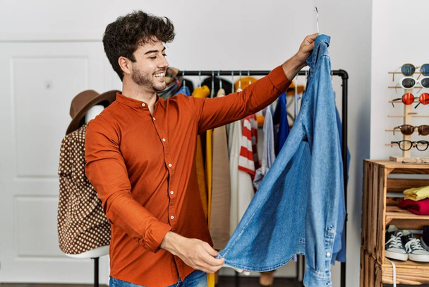 Joven cliente hispano sonriendo feliz sosteniendo percha con camisa en tienda de ropa. - Foto, imagen