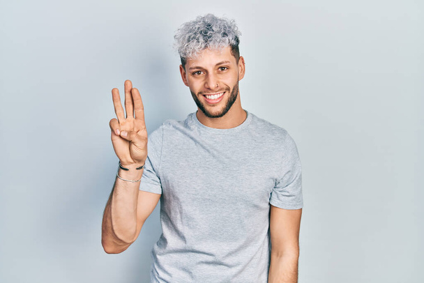 Joven hombre hispano con el pelo teñido moderno con camiseta gris casual mostrando y señalando con los dedos número tres mientras sonríe confiado y feliz.  - Foto, imagen