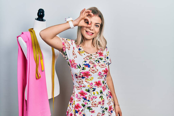 Beautiful caucasian woman dressmaker designer by manikin smiling happy doing ok sign with hand on eye looking through fingers  - Foto, Imagen