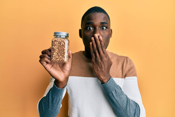 若いアフリカ系アメリカ人の男性が手で口をカバーレンズ豆の瓶を保持し、ショックを受け、間違いを恐れている。驚きの表情  - 写真・画像