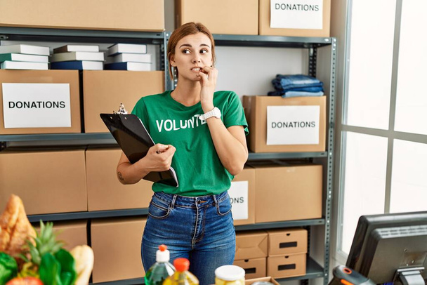 Młoda brunetka nosząca koszulkę wolontariuszki na darowiznach wygląda na zestresowaną i zdenerwowaną z rękami obgryzającymi paznokcie. niepokój.  - Zdjęcie, obraz