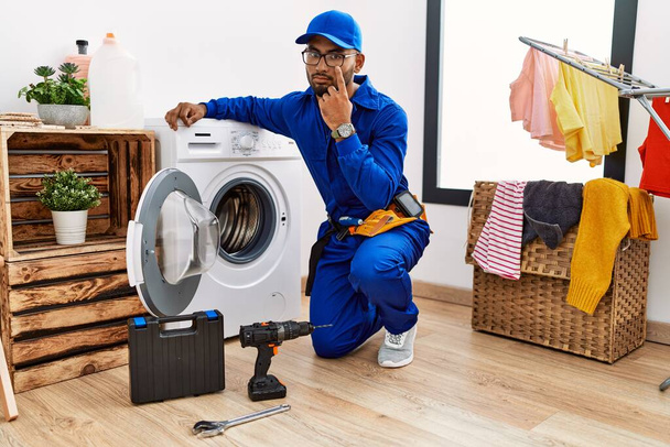 Çamaşır makinesinde çalışan genç Hintli teknisyen gözüne işaret ederek jestini ve şüpheli ifadeni izliyor.  - Fotoğraf, Görsel
