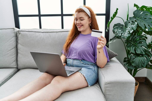 Jeune rousse femme souriant confiant en utilisant un ordinateur portable et une carte de crédit à la maison - Photo, image