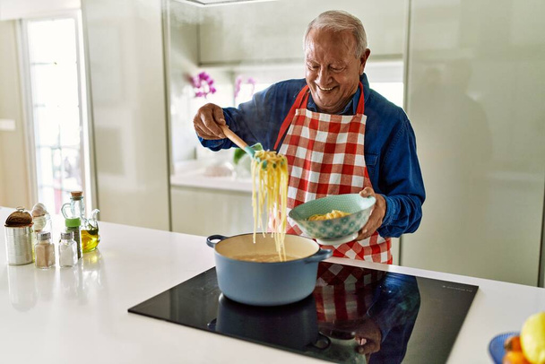 Senior man glimlacht zelfverzekerd koken spaghetti in de keuken - Foto, afbeelding