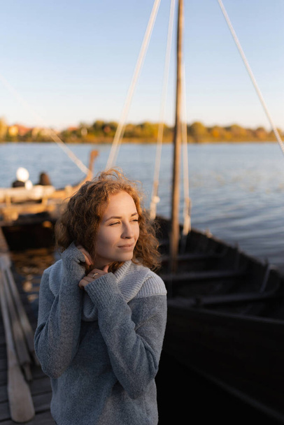 Genç kızıl saçlı kadın, kollarını başının yanında kaldırıyor, gülümsüyor ve gün batımını seyrediyor. Göl kenarında dinlen. - Fotoğraf, Görsel