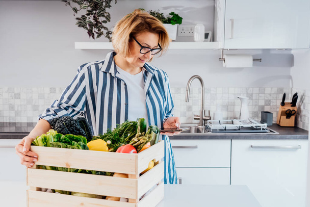 Online home food delivery. Woman checking her online order list on her phone. Wooden box with fresh vegetables and fruits standing on the kitchen table. Local farmer food. New Start of a healthy life. - Photo, Image