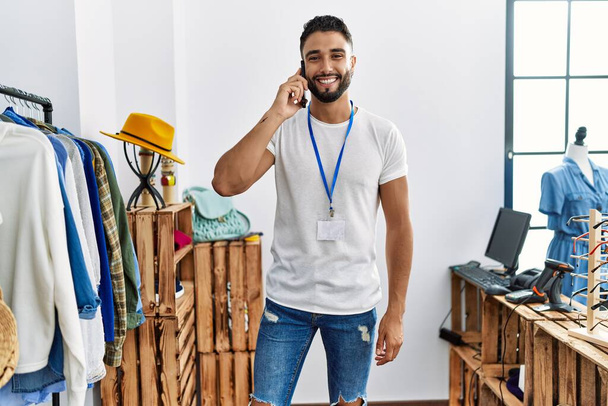 Junger arabischer Ladenbesitzer spricht auf dem Smartphone, das am Kleiderständer im Bekleidungsgeschäft steht - Foto, Bild