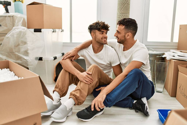 Два испаноязычных мужчины пара улыбается уверенно обнимая друг друга сидя в новом доме - Фото, изображение
