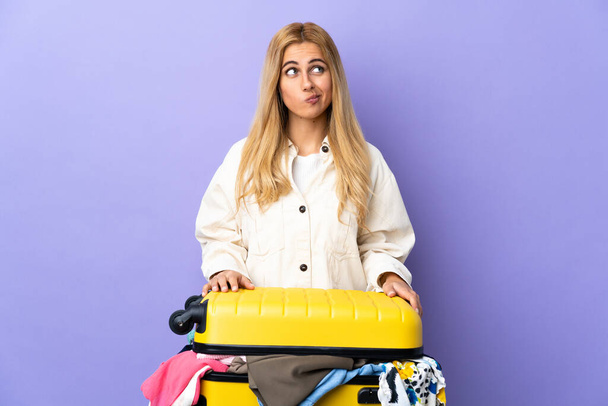 Young Uruguayan blonde woman with a suitcase full of clothes over isolated purple wall and looking up - Fotografie, Obrázek