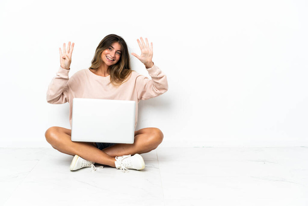 Jovem com um laptop sentado no chão isolado no fundo branco contando nove com os dedos - Foto, Imagem