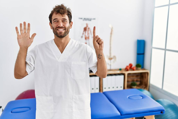 Jonge knappe fysiotherapeut werkt bij pijn herstel kliniek tonen en wijzen met de vingers nummer zes, terwijl glimlachen vol vertrouwen en gelukkig.  - Foto, afbeelding