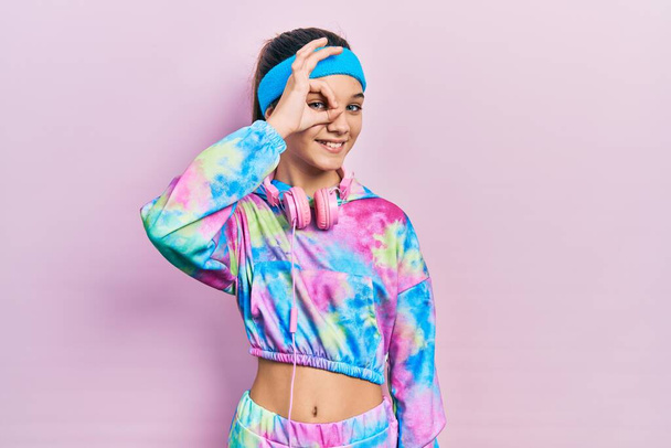 Young brunette girl wearing gym clothes and headphones smiling happy doing ok sign with hand on eye looking through fingers  - Фото, зображення
