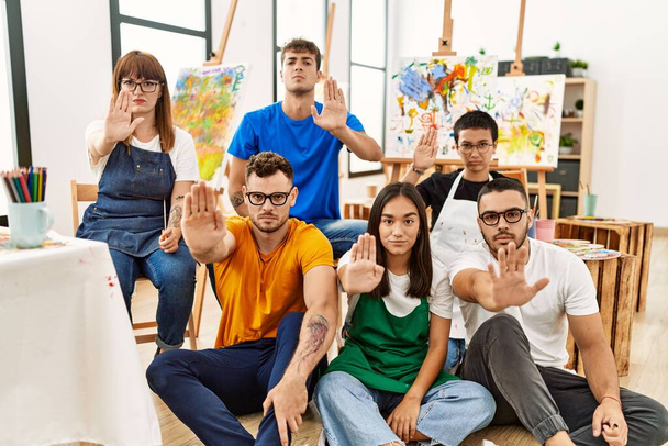 Grupo de personas sentadas en el estudio de arte con la mano abierta haciendo stop sign con expresión seria y segura, gesto de defensa  - Foto, imagen