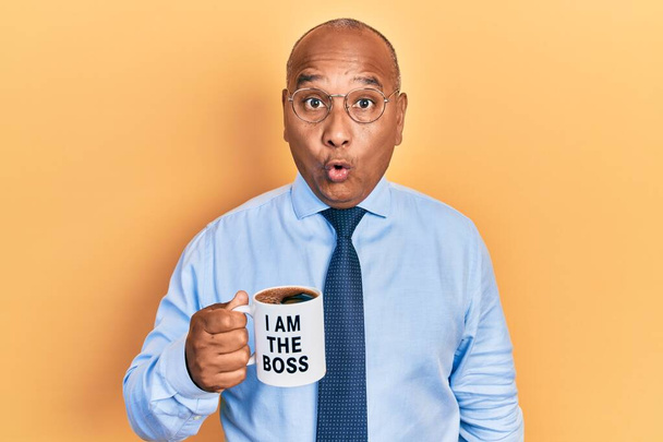 Hombre latino de mediana edad bebiendo de soy el jefe taza de café asustado y sorprendido con la boca abierta para sorpresa, cara de incredulidad  - Foto, imagen
