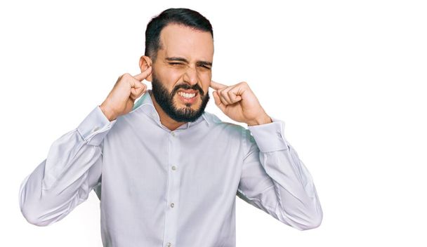 Egy szakállas fiatalember, aki üzleti inget visel, ujjakkal takarja el a fülét, bosszantó kifejezéssel a hangos zene zajára. süket fogalom.  - Fotó, kép