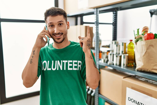 Jonge Spaanse man draagt vrijwillig t-shirt en spreekt aan de telefoon lachend vrolijk wijzend met hand en vinger naar de zijkant  - Foto, afbeelding