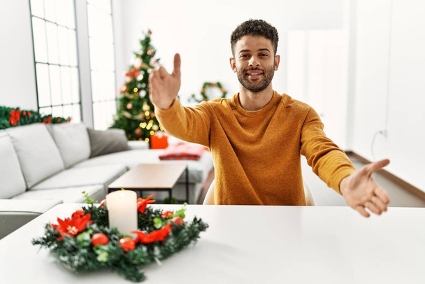 Arab young man sitting on the table by christmas tree looking at the camera smiling with open arms for hug. cheerful expression embracing happiness.  - 写真・画像