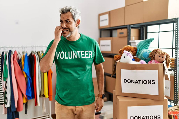 Латиноамериканец средних лет в футболке волонтёра на пожертвованиях выглядит напряжённым и нервным с руками во рту, грызущими ногти. тревожные проблемы.  - Фото, изображение