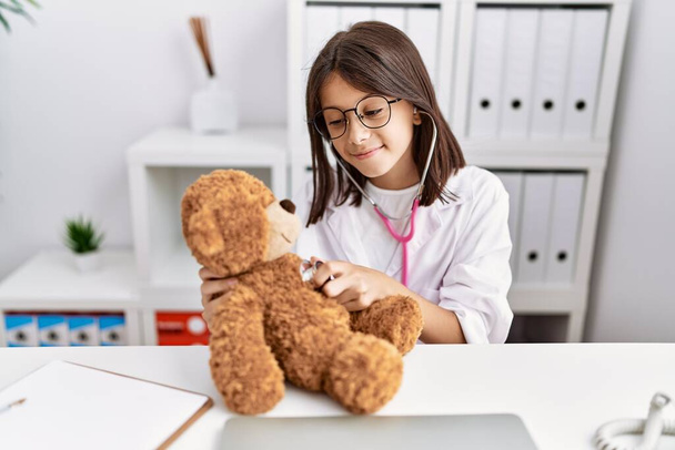 Joven chica hispana revisando la salud del oso de peluche en la clínica médica - Foto, imagen
