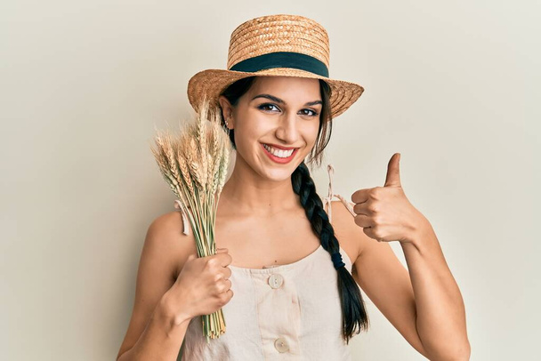 Молода іспаномовна жінка в літньому капелюсі тримає пряну пшеницю усміхненою щасливою і позитивною, великий палець робить відмінний знак схвалення
  - Фото, зображення