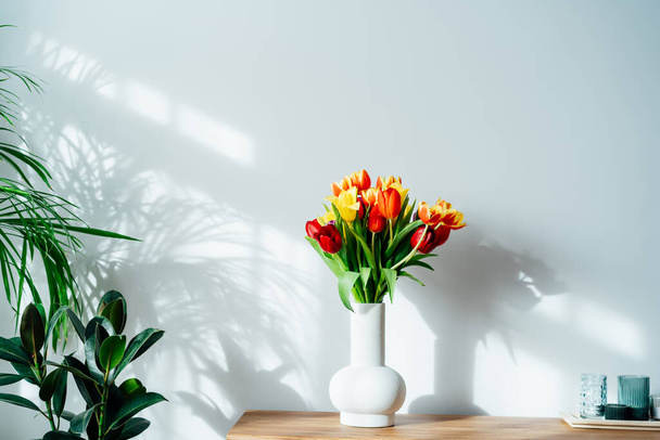 Scandinavian home interior with spring colorful bouquet of tulip flowers in ceramic vase standing on a wooden cabinet. Minimalist design with green plants and white wall. Biophilia style. Springtime. - Foto, Bild