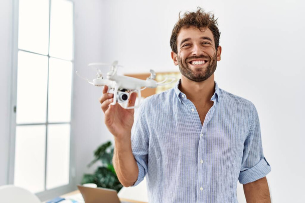 Νεαρός όμορφος άντρας που χρησιμοποιεί drone στο γραφείο του αρχιτέκτονα που δείχνει θετικός και χαρούμενος στέκεται και χαμογελάει με ένα σίγουρο χαμόγελο που δείχνει δόντια  - Φωτογραφία, εικόνα