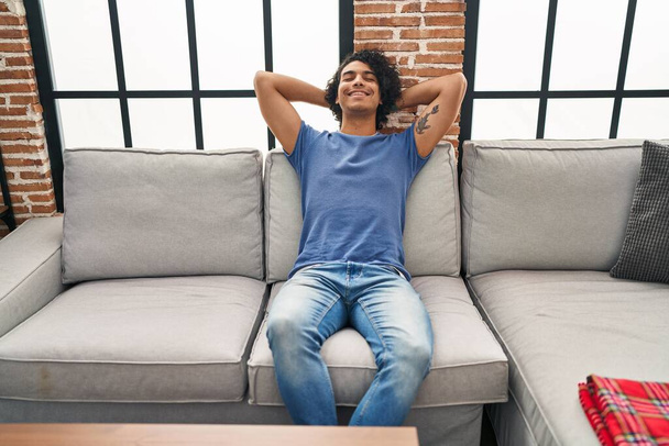 Giovane uomo ispanico rilassato con le mani sulla testa seduto sul divano a casa - Foto, immagini
