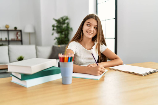 Чарівна дівчина робить домашнє завдання, сидячи на столі вдома
 - Фото, зображення