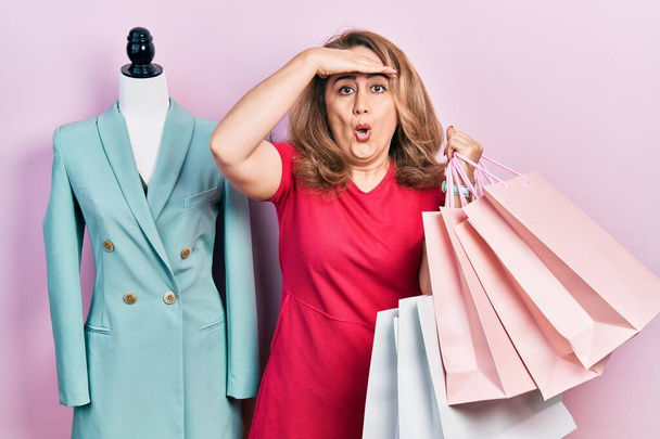 Mujer caucásica de mediana edad sosteniendo bolsas de compras estresadas y frustradas con la mano en la cabeza, cara sorprendida y enojada  - Foto, imagen