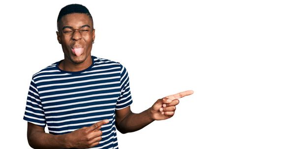 Junger afrikanisch-amerikanischer Mann zeigt mit den Fingern zur Seite und streckt die Zunge glücklich mit lustigem Gesichtsausdruck heraus.  - Foto, Bild