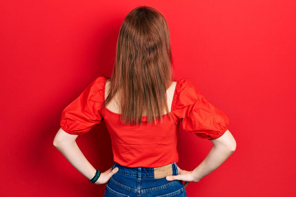 Junge rothaarige Frau in lässigem rotem T-Shirt, rückwärts stehend und mit den Armen am Körper wegschauend  - Foto, Bild