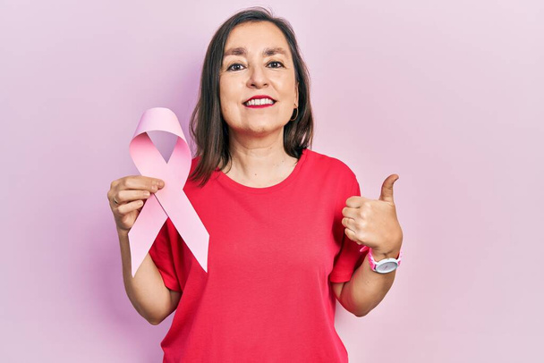 Middle age hispanic woman holding pink cancer ribbon smiling happy and positive, thumb up doing excellent and approval sign  - 写真・画像