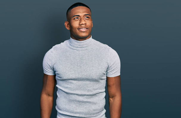 Jonge zwarte man draagt casual t-shirt kijken weg aan zij met glimlach op het gezicht, natuurlijke expressie. lachen vol vertrouwen.  - Foto, afbeelding
