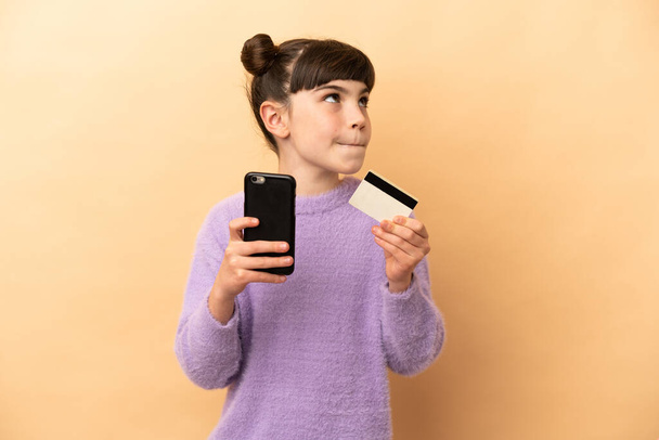Little caucasian girl isolated on beige background buying with the mobile with a credit card while thinking - Photo, Image