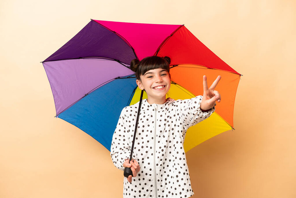 Holčička drží deštník izolovaný na béžovém pozadí s úsměvem a ukazující znamení vítězství - Fotografie, Obrázek