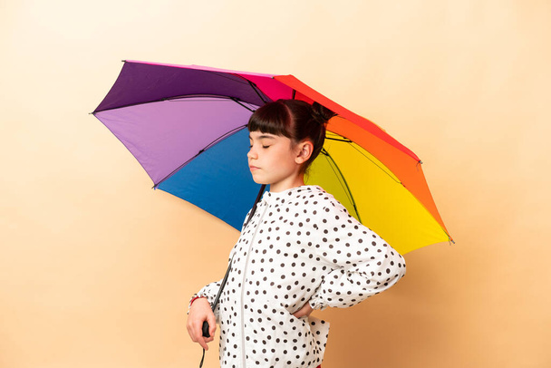 Μικρό κορίτσι που κρατά μια ομπρέλα απομονωμένη σε μπεζ φόντο που υποφέρει από πόνο στην πλάτη επειδή έκανε μια προσπάθεια - Φωτογραφία, εικόνα