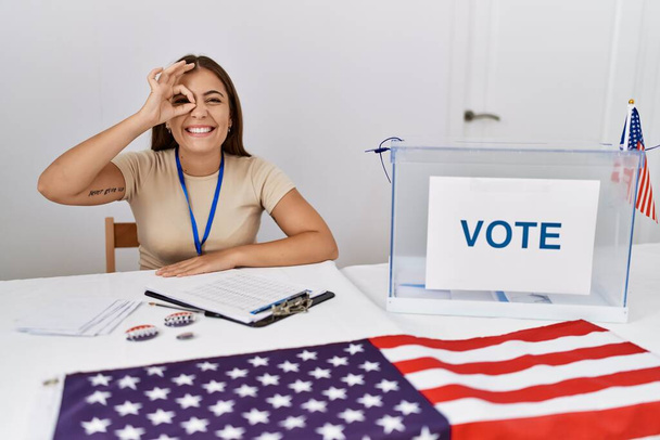 Jeune femme brune aux élections politiques assis par bulletin de vote souriant heureux faire ok signe avec la main sur les yeux regardant à travers les doigts  - Photo, image