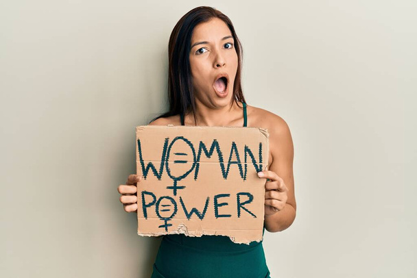 Junge lateinische Frau hält Frauenpower-Banner im Schockgesicht, sieht skeptisch und sarkastisch aus, überrascht mit offenem Mund  - Foto, Bild
