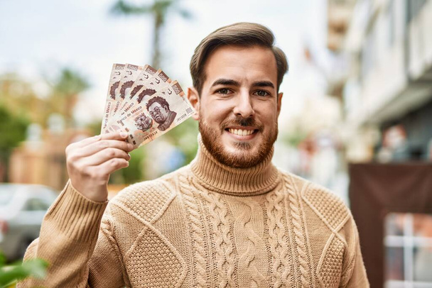 Fiatal fehér férfi mosolyog boldog gazdaság mexikói peso bankjegyek a városban. - Fotó, kép