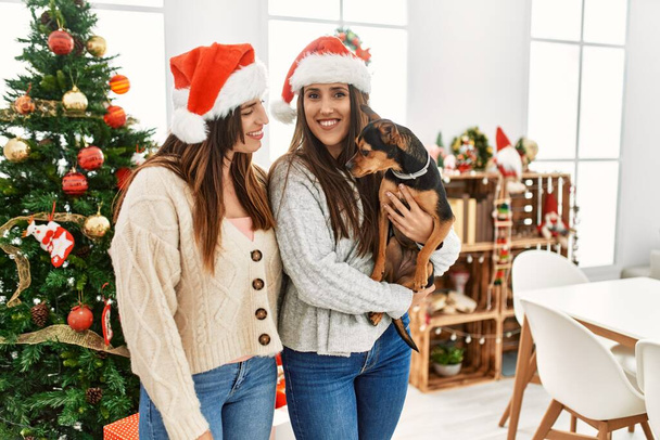 Two women smiling confident hugging dog standing by christmas tree at home - Photo, Image